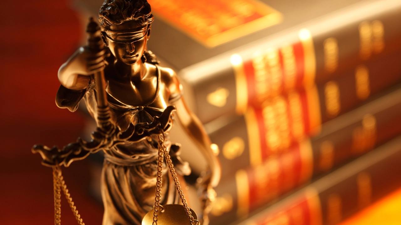 Justizia steht vor Bücherstapel mit Rechtstexten, sie hält eine Waage in der Hand und trägt eine Augenbinde